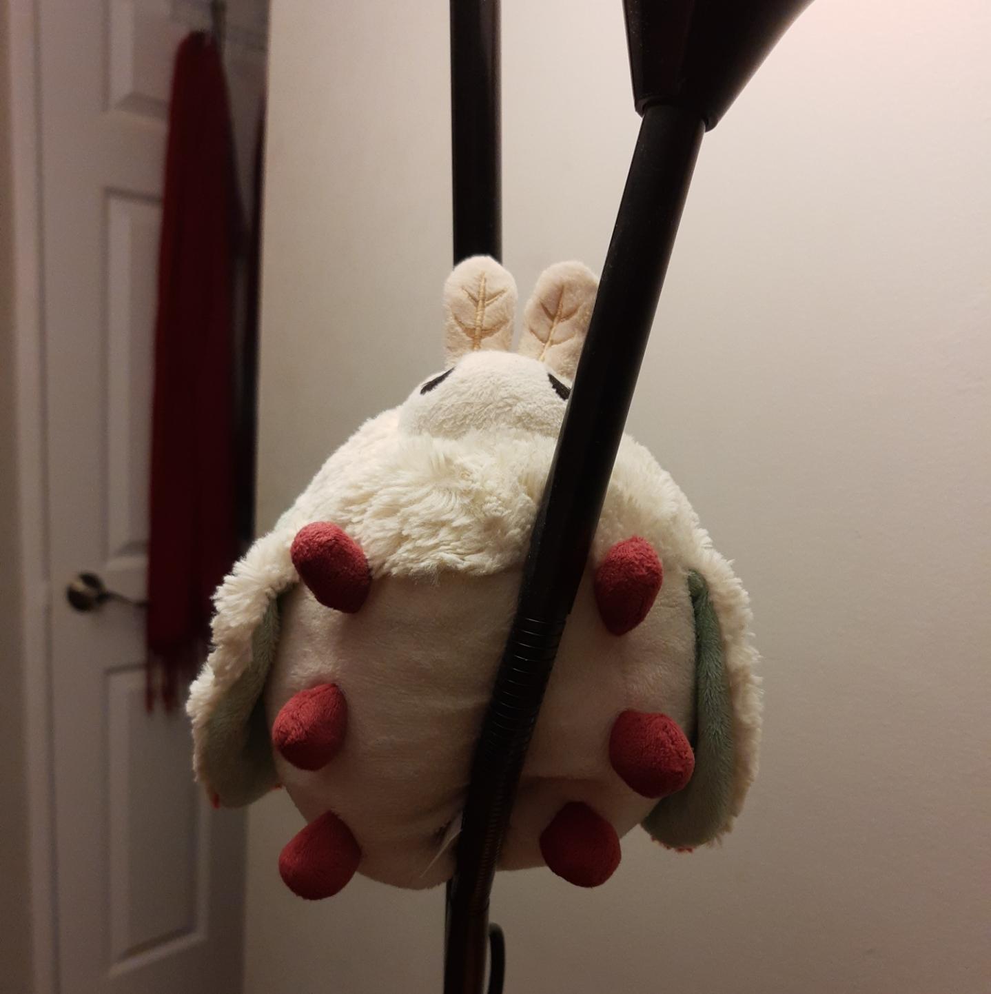 Photo of a white orb-like plushie moth with lil fuzzy attena and six lil magenta feet. It is wedged in a two light pole lamp in black with the bendy reading light arm pointing straight up. The way it is positioned, it looks like the lil moth legs are edging the pole so it can twirl.